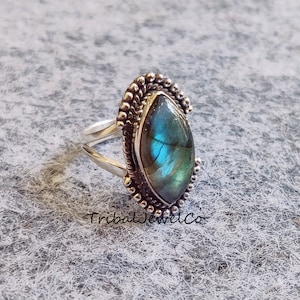 May include: A silver ring with a blue and green labradorite gemstone set in a detailed silver bezel. The ring has a beaded edge.