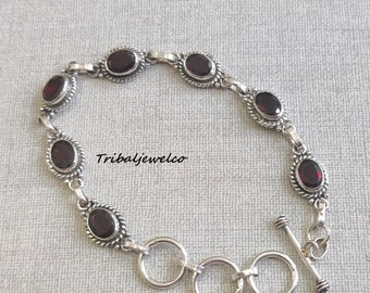 May include: A silver bracelet with a toggle clasp features a row of oval-shaped red gemstones. The bracelet is made of sterling silver and has a delicate design.