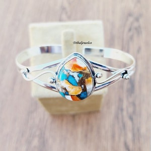 May include: A silver bangle bracelet with a teardrop-shaped turquoise and orange stone set in the center. The stone has a unique, marbled pattern of blue, orange, and white.