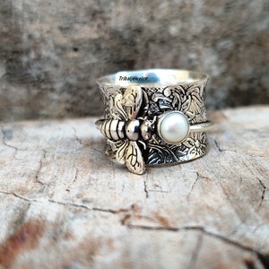 May include: A silver ring with a bee design and a pearl. The ring is engraved with floral patterns and the text "Tribaljewelco".