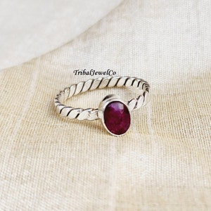 May include: A silver ring with a twisted band and a deep red oval gemstone. The ring is on a cream-colored fabric background.