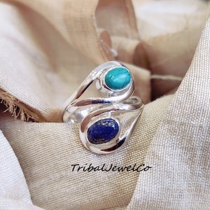 Puede incluir: Un anillo de plata con dos piedras ovaladas. La piedra superior es turquesa y la piedra inferior es lapislázuli. El anillo está sobre un fondo de tela beige. 'TribalJewelCo' está impreso en la tela.