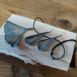 Op de afbeelding: Een koperkleurige sieradenset met een hanger en oorbellen, elk in de vorm van een gestileerd blad. De hanger hangt aan een zwart koord en de oorbellen hebben haaksluitingen. De set ligt op een houten oppervlak.