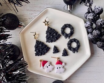 May include: A collection of black Christmas-themed earrings displayed on a white plate. The earrings include Christmas trees, wreaths, and ghost-like figures wearing Santa hats. Gold star accents are visible on some earrings. Black ornaments and tinsel surround the plate.