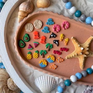 Op de afbeelding: Een verzameling miniatuur, kleurrijke strandthema bedels en decoraties. Inclusief avocado's, watermeloenen, ananassen, octopussen, garnalen, zeepaardjes, zeesterren en meer. De bedels zijn gerangschikt op een terracotta-kleurige tray met zand en schelpen.