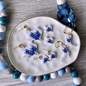 Puede incluir: Un conjunto de seis pares de pendientes que presentan un contorno blanco del estado de Texas con acentos de flores azules. Los pendientes se muestran en un plato blanco con un borde dorado.