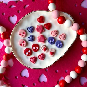 Könnte beinhalten: Ein weißer, ovaler Teller mit verschiedenen Verzierungen zum Valentinstag. Der Teller enthält rote, rosa und lila herz- und schädelförmige Dekorationen. Eine Perlenkette mit roten, weißen und rosa Perlen umgibt den Teller, vor einem rosa Hintergrund.