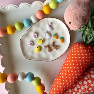 May include: A white ceramic dish with a gold rim holds a variety of small Easter-themed charms. The charms include bunnies, chicks, carrots, and footprints. The dish is surrounded by a colorful garland of wooden beads.
