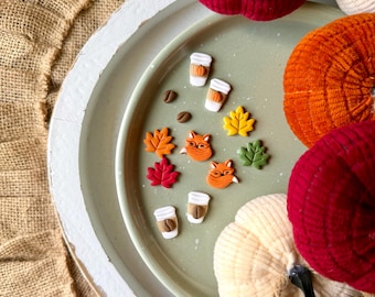 Könnte beinhalten: Eine Sammlung herbstlicher Dekorationsartikel. Enthält Miniaturkürbisse in Rot, Orange und Creme, zusammen mit kleinen Verzierungen von Kaffeetassen, Fuchsköpfen und Herbstblättern. Die Artikel sind auf einem grünen Tablett angeordnet.