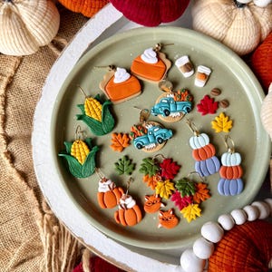 May include: A selection of autumn-themed earrings, featuring pumpkin pie slices, corn, coffee cups, and a blue truck laden with pumpkins. The earrings are in shades of orange, green, and blue, displayed on a green plate.