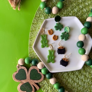 Puede incluir: Un conjunto de cuatro pares de pendientes con un trébol de cuatro hojas, una olla de oro, una herradura y un osito de goma. Los pendientes se muestran en un plato blanco con un fondo verde.