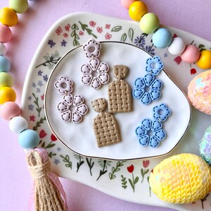 May include: A collection of handmade earrings in various designs, including floral and woven patterns. The earrings are in shades of pink, blue, and tan, displayed on a floral-decorated plate. A colorful beaded garland and sequined eggs add to the decorative display.