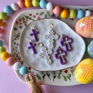 May include: A collection of earrings in a variety of cross designs. Some are solid purple, others are outlined in gold, and some are adorned with white daisy-like flowers. The earrings are displayed on a floral-patterned plate with colorful beads and sequined Easter eggs.