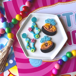 May include: Two pairs of polymer clay earrings on a white hexagonal tray. One pair features light blue abstract shapes with small green circles and pink peppermint-like accents. The other pair depicts black and yellow bumper cars with blue smoke.
