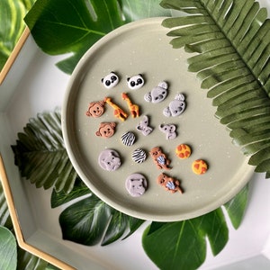 Puede incluir: Una colección de 14 pequeños pendientes de animales coloridos en un plato verde claro. Los pendientes están hechos de arcilla polimérica y presentan una variedad de animales, incluidos pandas, koalas, monos, jirafas, elefantes y nutrias.