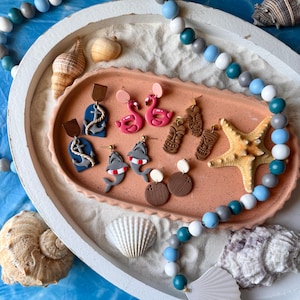 May include: A collection of handmade earrings with a beach theme. The earrings feature designs such as anchors, flamingos, tiki masks, and sharks. The earrings are displayed on a pink tray with sand, seashells, and a beaded necklace.
