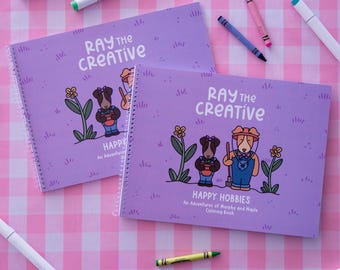 May include: Two spiral-bound coloring books with light purple covers featuring the text Ray the Creative Happy Hobbies An Adventures of Murphy and Maple Coloring Book, illustrated with two cartoon dogs, flowers, and grass, surrounded by markers and crayons.