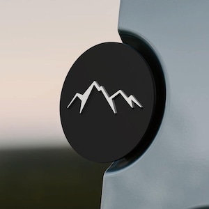 May include: A black, circular emblem featuring a white mountain range design. The emblem is affixed to a light-colored surface, creating a contrast between the dark and light elements. The mountain range is a simple, stylized graphic.
