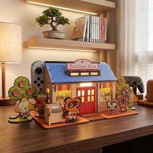 Könnte beinhalten: Ein Holzmodell des "Boutique Nook"-Ladens aus Animal Crossing, mit blauem Dach und roten Türen. Das Modell ist beleuchtet und enthält Charakterfiguren. Ein Nintendo Switch-Controller ist im Hintergrund sichtbar.