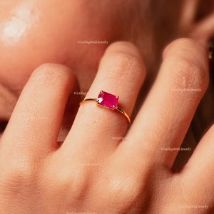 Puede incluir: Un anillo de oro con una piedra rectangular de color rosa brillante. El anillo está en el dedo de una persona.