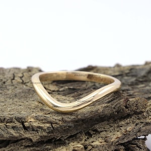 May include: A simple gold band ring with a curved, wave-like design. The ring is shown on a piece of wood.