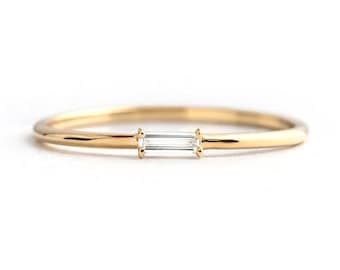 May include: A delicate gold ring with a thin band and a rectangular, clear gemstone set horizontally in the center. The ring has a minimalist design, perfect for everyday wear. The ring is set against a white background.