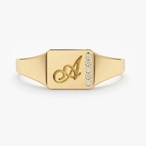 May include: A gold signet ring with a square face featuring a cursive letter 'A' and a row of small diamonds down the side.