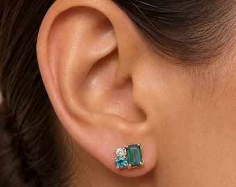 May include: A gold stud earring featuring a cluster of three gemstones: a rectangular emerald-cut green stone, a round brilliant-cut clear stone, and a smaller round-cut bright blue stone, all set in a polished gold metal base on an earlobe.