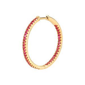 Puede incluir: Un pendiente de aro dorado con una fila continua de pequeñas gemas de color rosa brillante engastadas a lo largo del borde exterior. El pendiente se muestra sobre un fondo blanco liso, resaltando su diseño y color.