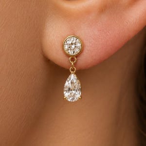 May include: A gold drop earring featuring a round bezel-set clear stone at the top connected by a small jump ring to a pear-shaped clear stone at the bottom. The earring is crafted from polished yellow gold metal and displays two faceted stones.