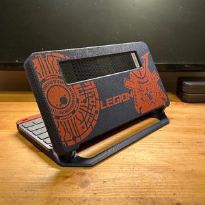 May include: A dark blue handheld gaming device with a textured finish, featuring orange graphic prints of a circuit pattern and a stylised helmet. The word LEGION is printed in orange on the centre, with a black vent and a keyboard visible below.