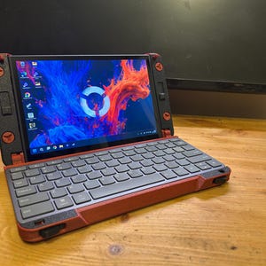 Puede incluir: Un portátil pequeño y abierto, con un teclado negro y una carcasa roja y negra. La pantalla muestra un diseño abstracto colorido. El portátil está sobre un escritorio de madera, con un monitor y un portalápices en el fondo.