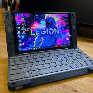 Puede incluir: Un ordenador portátil negro y gris con una pantalla que muestra un gráfico colorido con temática espacial y la palabra "LEGION". El teclado es gris con letras blancas. El portátil está abierto sobre una superficie de madera.