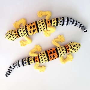 May include: Two crocheted gecko lizards with yellow, orange, black, and white stripes. The geckos are lying on a white surface.