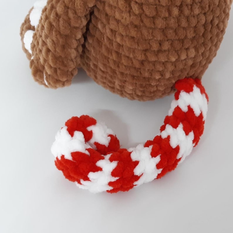 May include: A close-up of a brown and white crocheted stuffed animal. The body is a solid brown color, and the tail is red and white striped. The toy is made of a soft, plush material.