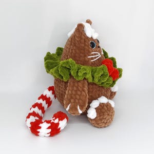 May include: A handmade brown crochet cat plushie with a white and red candy cane tail. The cat has a green wreath around its neck with red berries and white accents. The cat's eyes are blue.