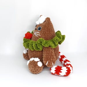 May include: A brown plush cat with blue eyes, white whiskers, and a white hat. It has a green collar, a red flower, and a red and white striped tail. The cat is sitting on a white surface.