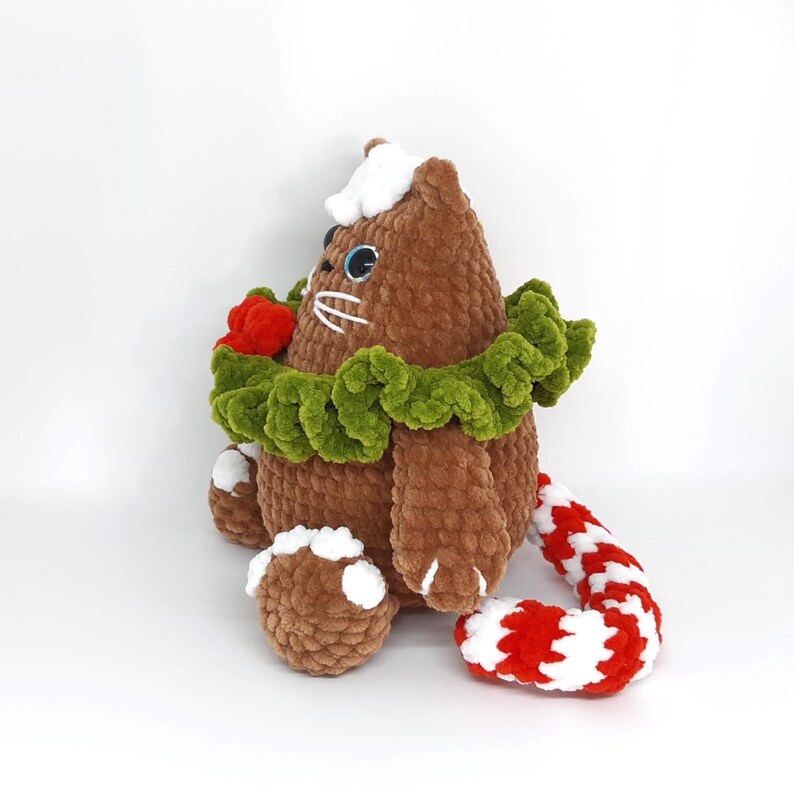 May include: A brown plush cat with blue eyes, white whiskers, and a white hat. It has a green collar, a red flower, and a red and white striped tail. The cat is sitting on a white surface.