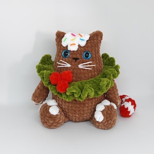 May include: A brown crocheted cat plushie with blue eyes, white whiskers, and a green wreath around its neck. The cat has a white frosting-like topping with colorful sprinkles and white accents on its paws. A red and white candy cane is next to the cat.