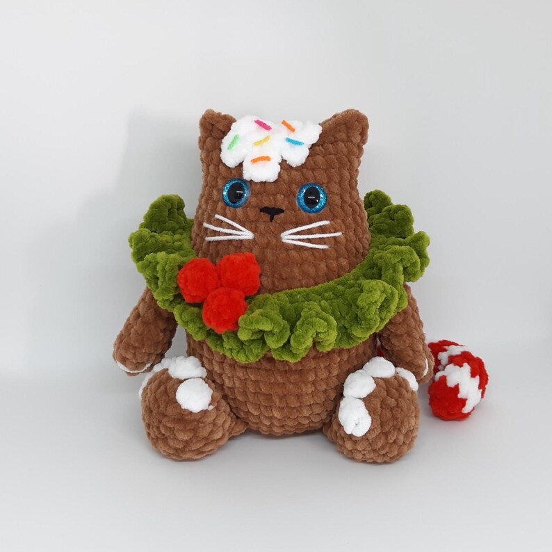 May include: A brown crocheted cat plushie with blue eyes, white whiskers, and a green wreath around its neck. The cat has a white frosting-like topping with colorful sprinkles and white accents on its paws. A red and white candy cane is next to the cat.