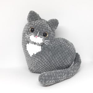 May include: A gray plush cat toy with a white chest and whiskers. The cat has large, round eyes with gold accents and a pink nose. The tail is curled. The toy is made of a soft, textured material.