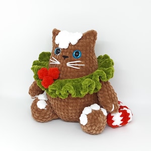 May include: A handmade brown crochet cat plushie with white whiskers, blue eyes, and a green ruffled collar adorned with red berries. The cat has white accents on its head and paws, and a red and white striped candy cane.