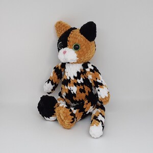 May include: A crocheted calico cat plush toy with black, white, and orange fur. The cat has green eyes and a pink nose.