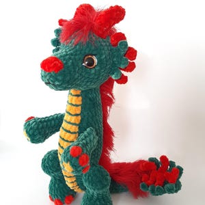 May include: A handmade plush dragon toy in shades of green, red, and yellow. The dragon features a textured body, with red accents on its head, tail, and feet. The dragon has large, round eyes and a friendly expression.