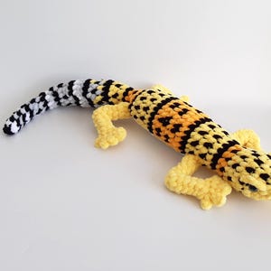 May include: A handmade crochet gecko toy in yellow, black, and orange. The gecko has a long tail with black and white stripes, a body with a spotted pattern, and yellow feet. The toy is lying on a white surface.