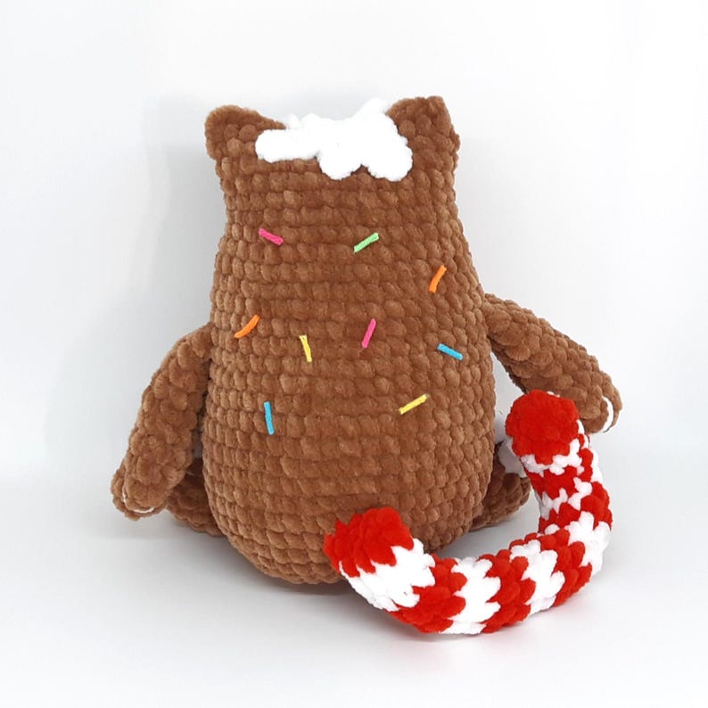 May include: A brown crocheted plush toy shaped like a bear with white frosting and colorful sprinkles. It has small arms and is holding a red and white striped candy cane. The toy is on a white background.