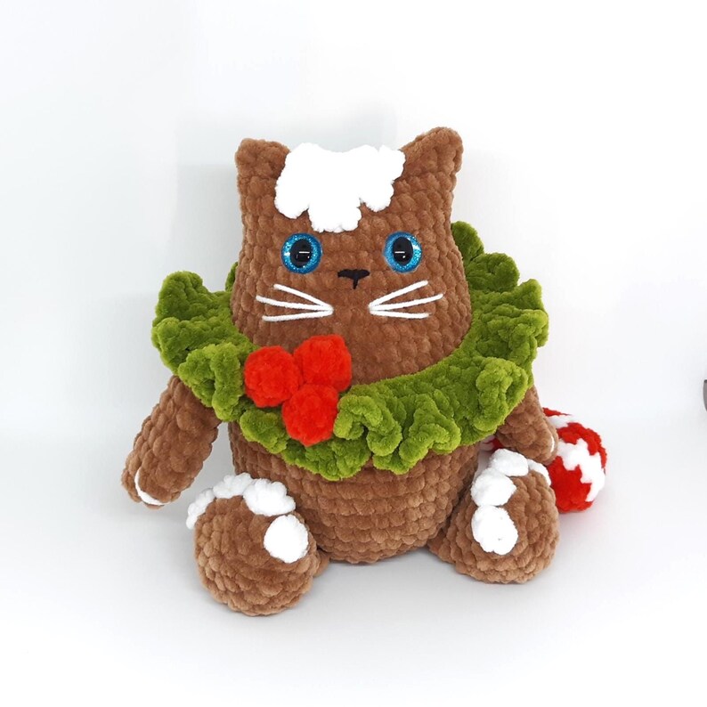 May include: A brown crocheted cat plush toy with blue eyes, white whiskers, and a white patch on its head. It wears a green ruffled collar with red berry accents and has white and red accents on its paws and tail.