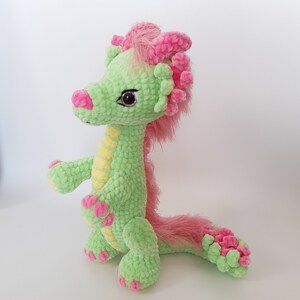 May include: A green crocheted dragon with pink accents and a fluffy pink tail. The dragon has yellow stripes down its back and large, black eyes.