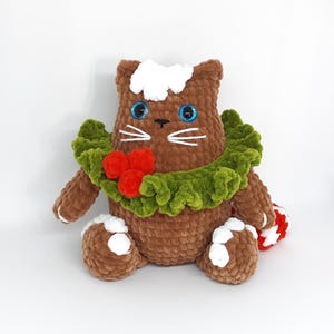 May include: A handmade brown crochet cat plushie with blue eyes, white whiskers, and a green wreath with red berries. The cat has white accents on its paws and head, and holds a red and white striped candy cane.