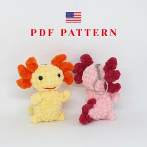 Peut inclure: Deux porte-clés axolotl crochetés. L'un est jaune avec des branchies orange, l'autre est rose avec des branchies bordeaux. Les deux ont des yeux noirs et des sourires rouges. Le texte "PDF PATTERN" est au-dessus des axolotls, avec le drapeau américain en haut à gauche.